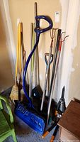 Assorted garden and yard tools including rakes, brooms, snow shovel with blue handle, and plastic garbage bin.