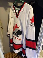 White Nike Team Canada hockey jersey with red and black stripes and maple leaf logos on front and sleeves.