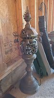 Full view of ornate brass outdoor lantern light fixture with base and top finial against plywood wall