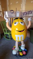 Front view of full display showing yellow M&M character holding transparent bin atop head, round black base with molded M&M spheres and visible wheels