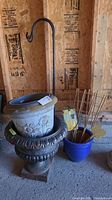 Group shot of cast iron planter, beige plastic planter, blue pot with bamboo supports, and shepherd’s hook against plywood wall