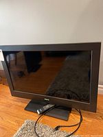 Full view of the 32 inch LCD RCA TV powered off on wooden floor with remote placed on base stand.