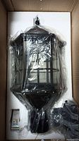 Box containing a black framed ball lamp with clear side panels, packaged with protective foam and plastic wrap.