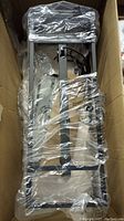 Rectangular metal framed light fixture wrapped in plastic inside cardboard box.