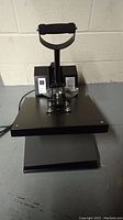 Front view of a black heat press machine showing the flat pressing surface, handle, and control box.