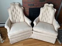 Photo showing the front view of two cream-colored tufted armchairs with scalloped back and rolled arms, in used condition with fabric wear and some marks.
