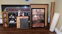 Photo showing framed Rocky Marcia boxing artwork, AEW wrestling poster, multiple framed portraits and art, plus some empty or small frames.