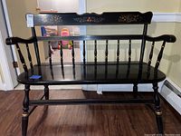 Front view of a black wooden bench with turned legs and spindles, gold accents, and floral painting on the backrest.