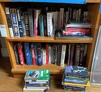 Wooden bookshelf with two shelves filled with hardcover and paperback books covering sports history, biographies, and fiction. Stacked VHS tapes in front of the shelf.