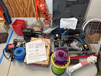 Wide view showing all items on a table and hanging on a wooden board including the Ryobi drill case open with drill visible, handheld vacuum, sander, sanding paper, red container, and extension cord.