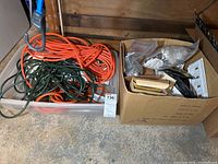 Plastic bin with orange and green extension cords on left, cardboard box with electrical outlets, switches, faceplates, and wiring components on right