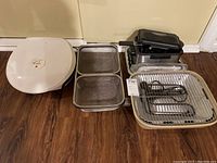 Lot setup showing all items in lot together on wooden floor: George Foreman grill, two metal mesh baskets, compact fryer, electric grill base with cooking coils and rack.