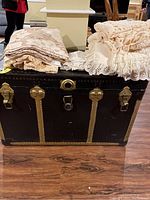 Folded assorted lace and crochet linens placed on a black storage trunk, showing different patterns and textures including floral lace and fringed crochet
