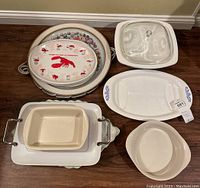 Photo of assorted ceramic and plastic platters, casserole dishes, and bakeware pieces on floor with wood grain surface.