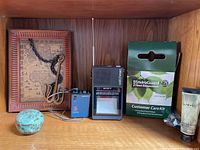 Vintage Sony Walkman and Sony Watchman portable TV on wooden shelf alongside framed map with chain and key, EnviroGuard NG Care Kit box, small pottery, and tubes.