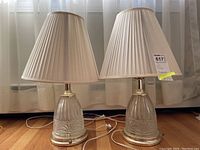 Pair of glass base table lamps with metal accents and pleated fabric lampshades on wood floor in front of white curtain