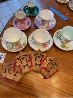 Six gold-rimmed bone china teacups and saucers on a wooden table with wood carved bowl server tied with red fabric.