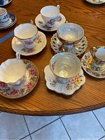Photo of assorted bone china teacups and saucers on wooden table showing variety in shape, pattern and gold rim detail