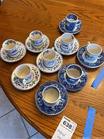 Overall view of nine espresso teacups and saucers in two distinct patterns arranged on a wooden table with a sale tag and blue tape for scale.