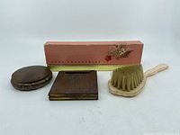 Photo shows two compacts, a hair or makeup brush, and a pink Avon fragrance box with floral design arranged in a row.