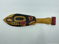 Front view of the carved and painted wooden paddle showing the native indigenous design in black, red and gold on the blade and red painted end of the handle.