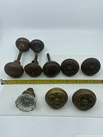 Front and side view of seven metal door knobs, one glass door knob, and one smaller metal knob, with a tape measure showing size.