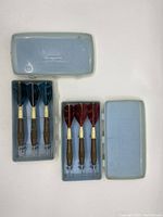 Two open plastic cases each containing three used darts with feather flights; one set blue, one set red flights