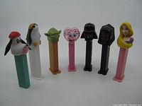 Front view of 7 unpackaged PEZ dispensers featuring vintage and Star Wars character heads including dog, penguin, Yoda, heart face, Darth Vader, Maleficent, and blonde female character.
