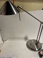 Full view of silver metal desk lamp showing conical shade, adjustable arm, and base on white surface.
