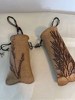 Pair of textured ceramic clay pouches with botanical designs and black cords on a white surface.