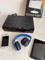Overview of electronics items: DVR recorder, Samsung Galaxy 5 smartphone, blue Beats headphones, Amazon Kindle, Apple AirPort Extreme router box