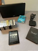 Overview photo showing all items including Ion Bluetooth speaker, cassette tapes, boxed FitSport Bluetooth earphones, Amazon Kindle, Apple TV device with cable, and video editing software DVD case.