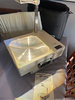 Front angled view of Elmo HP-285p overhead projector on a table, showing projector glass surface, handle, and part of the metallic arm. Instruction manual laid flat in front and carry bag in background.