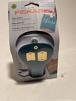 Front view of Fiskars tag maker punch in original packaging, showing branding, product name, and sample tags.