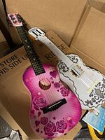 Two children's guitars inside a box; one pink with rose and heart design, one white with gray decorative patterns