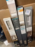 Four boxed vinyl mini blinds in various sizes stacked inside a cardboard box. Three boxes visible with size labels 35x64 in, 31x64 in, others not fully visible.