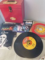 Display of red vintage record storage case with a variety of 45 RPM records and sleeves including Queen, Elvis Presley, Pablo Cruise.