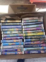 Top-down view of two layers of Disney VHS tapes stacked in a banana box showing a variety of titles and colorful covers
