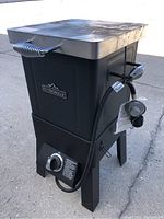 Front side view of the RiverGrille propane deep fryer, showing black body, control knob, side spring handles, and hose with regulator attached.