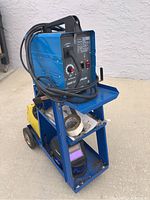 Blue Chicago Electric welder unit on top shelf of blue metal cart with cables wrapped around.