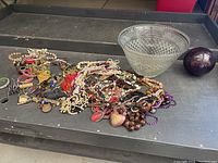 Overview of mixed lot showing crystal punch bowl, large dark red orb, and assortment of costume jewelry with various colors and styles.