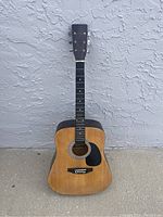 Front view of old acoustic guitar with natural wood finish and black pickguard against textured wall.