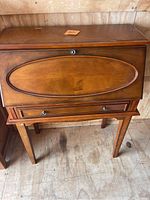 Front view of the wooden secretary desk with the fold-down front panel closed, showing an oval recessed design and a knob.