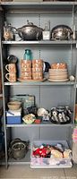 Shelf with multiple items including stacks of stoneware cups and plates, silver plated chafing dishes, assorted kitchenware and collectibles