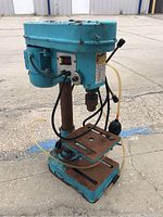 Front angled view of vintage blue drill press showing power switch, motor housing, and rust on base and table.