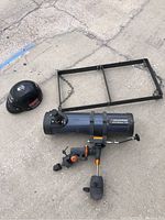 Three items on pavement: a Celestron AstroMaster 130 telescope with mount, black cast iron hanging rack with chains, and a black motorcycle helmet with stickers.