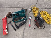 Wide view showing a variety of hand tools including hammer, adjustable wrench, various small tools in a green toolbox, power tools including a saw, vice, and two extension cords coiled up.