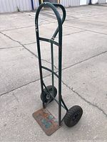 Full view of two wheel dolly showing green metal frame, round handle, rusted base plate, and black tires.