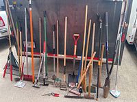 Full view of all yard tools lined against a wall including rakes, shovels, sledgehammer, and other implements.