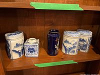 Five Asian-style ceramic items on wooden shelf, comprising three jars with lids and two vases in blue and white and cobalt blue colors.